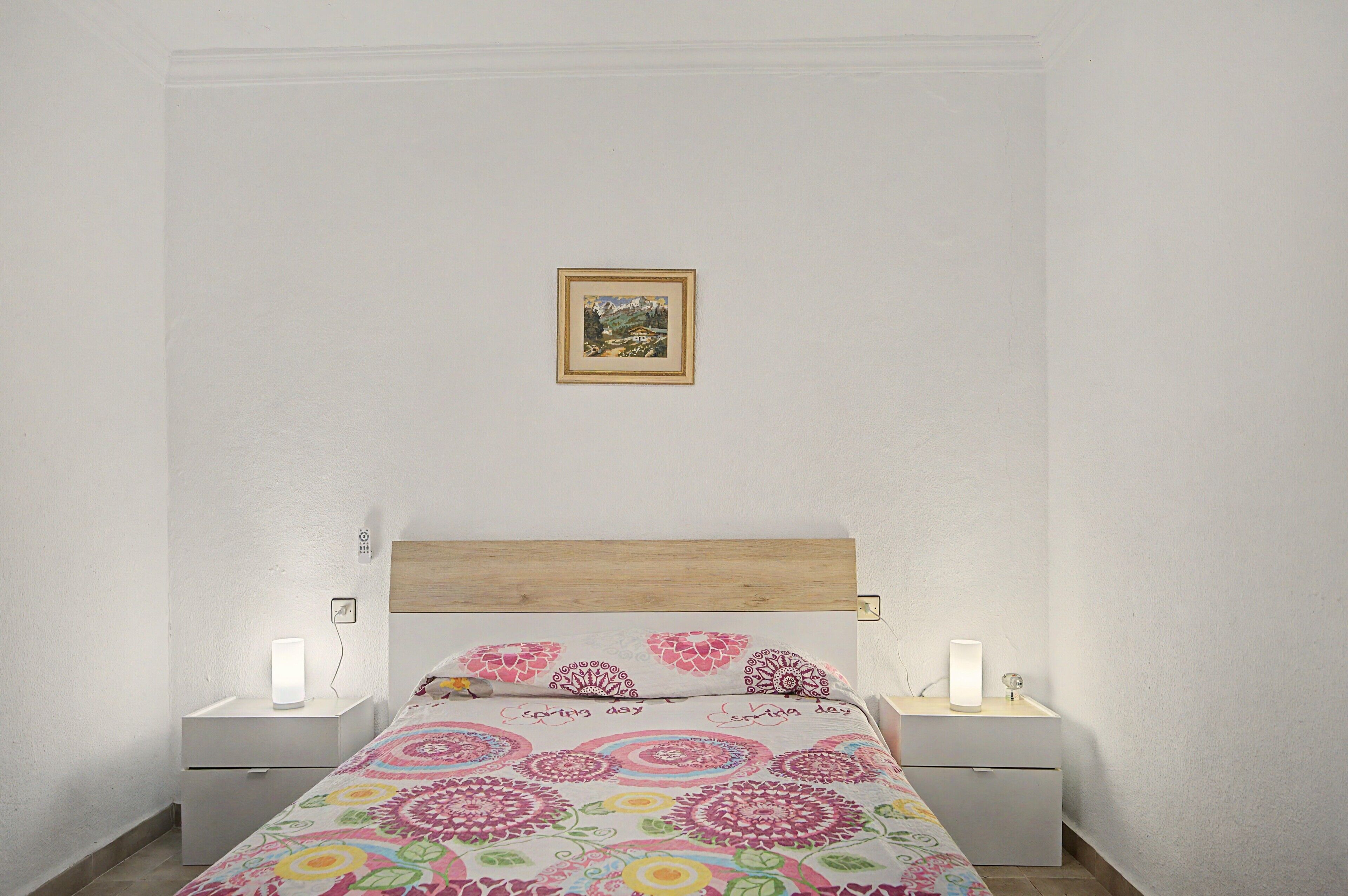 Un dormitorio con cama, dos mesitas de noche, lámpara de noche y un cuadro en la pared.