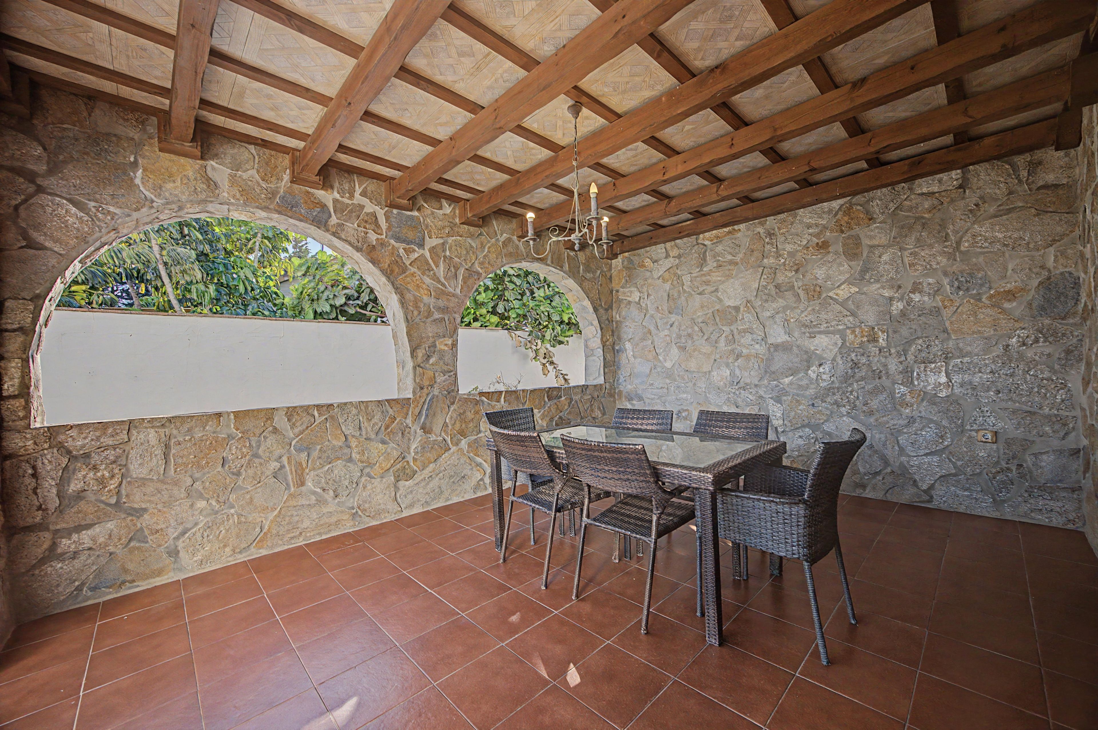 Una stanza con pareti in pietra, pavimento piastrellato, travi in legno a soffitto, un tavolo in vetro con sedie in vimini e due finestre ad arco con piante.