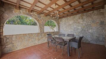 Una stanza con pareti in pietra, pavimento piastrellato, travi in legno a soffitto, un tavolo in vetro con sedie in vimini e due finestre ad arco con piante.