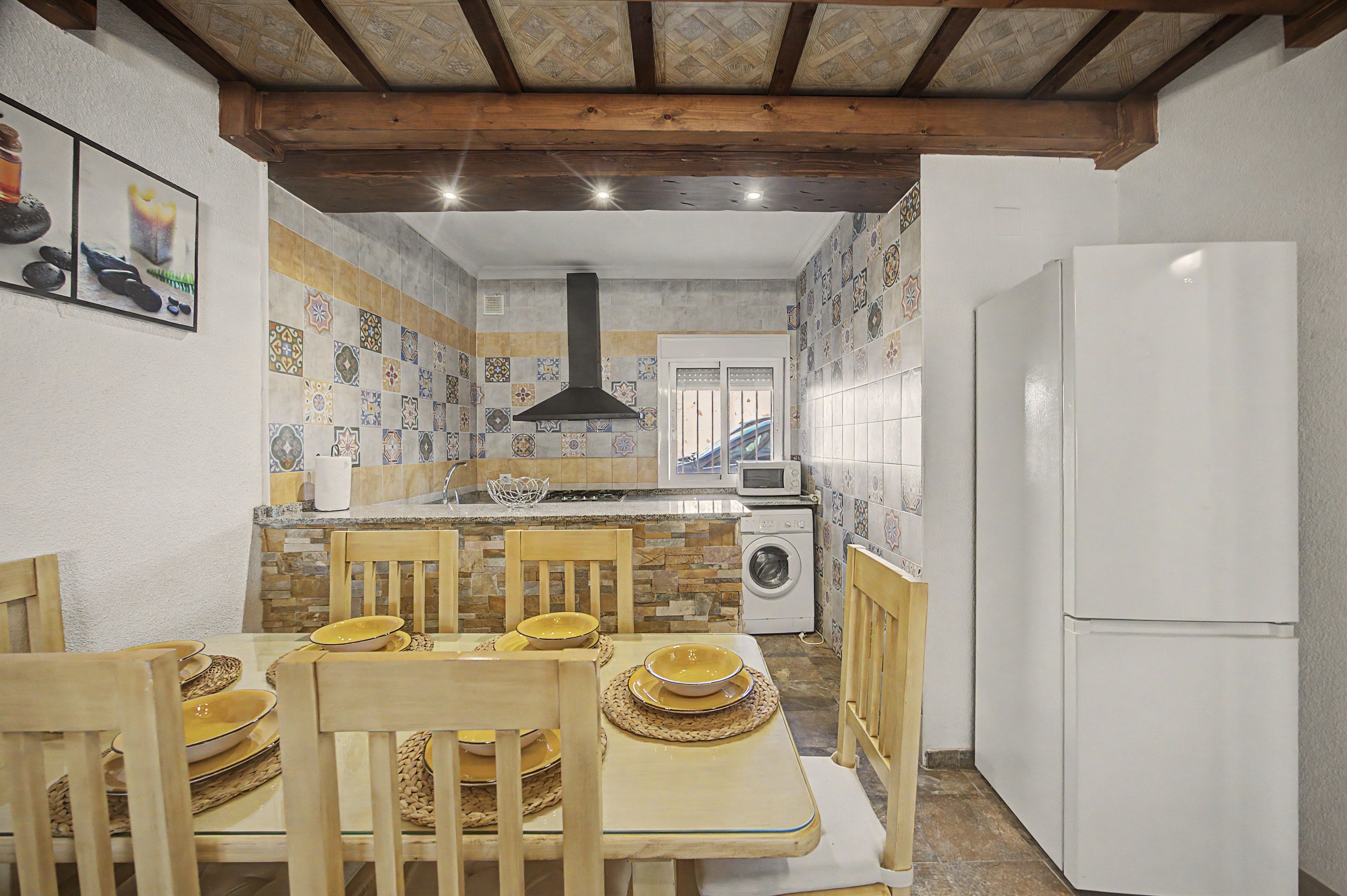 Cocina compacta con comedor, que incluye una isla central, sillas de madera, lavadora integrada y refrigerador blanco.