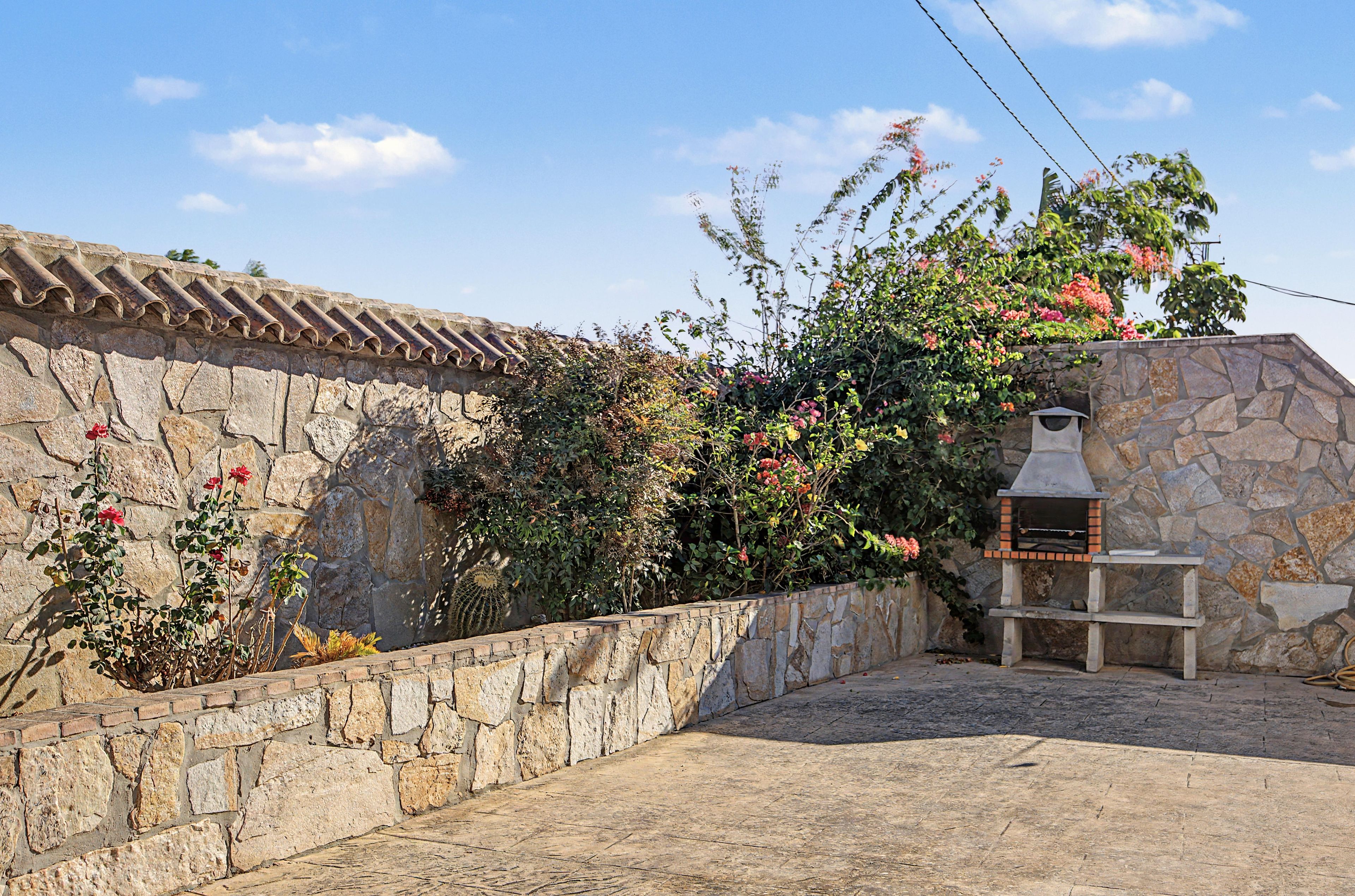 Un muro de piedra con techo de tejas, una parrilla de piedra y una variedad de plantas con flores.