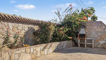 Un muro de piedra con techo de tejas, una parrilla de piedra y una variedad de plantas con flores.