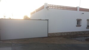 A building with a closed white gate, a stone base, and a paved area with a circular pattern. - Holiday Home 'Casa El Arriate' with Shared Pool and Private Terrace (Vejer de la Frontera)