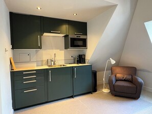 Een moderne keuken met donkere kastjes, een magnetron en een spoelbak. In de hoek staat een bruine fauteuil en een vloerlamp.