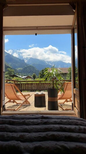 Un balcón con sillas de madera y una planta en maceta, con vista a un paisaje montañoso y cielo despejado.