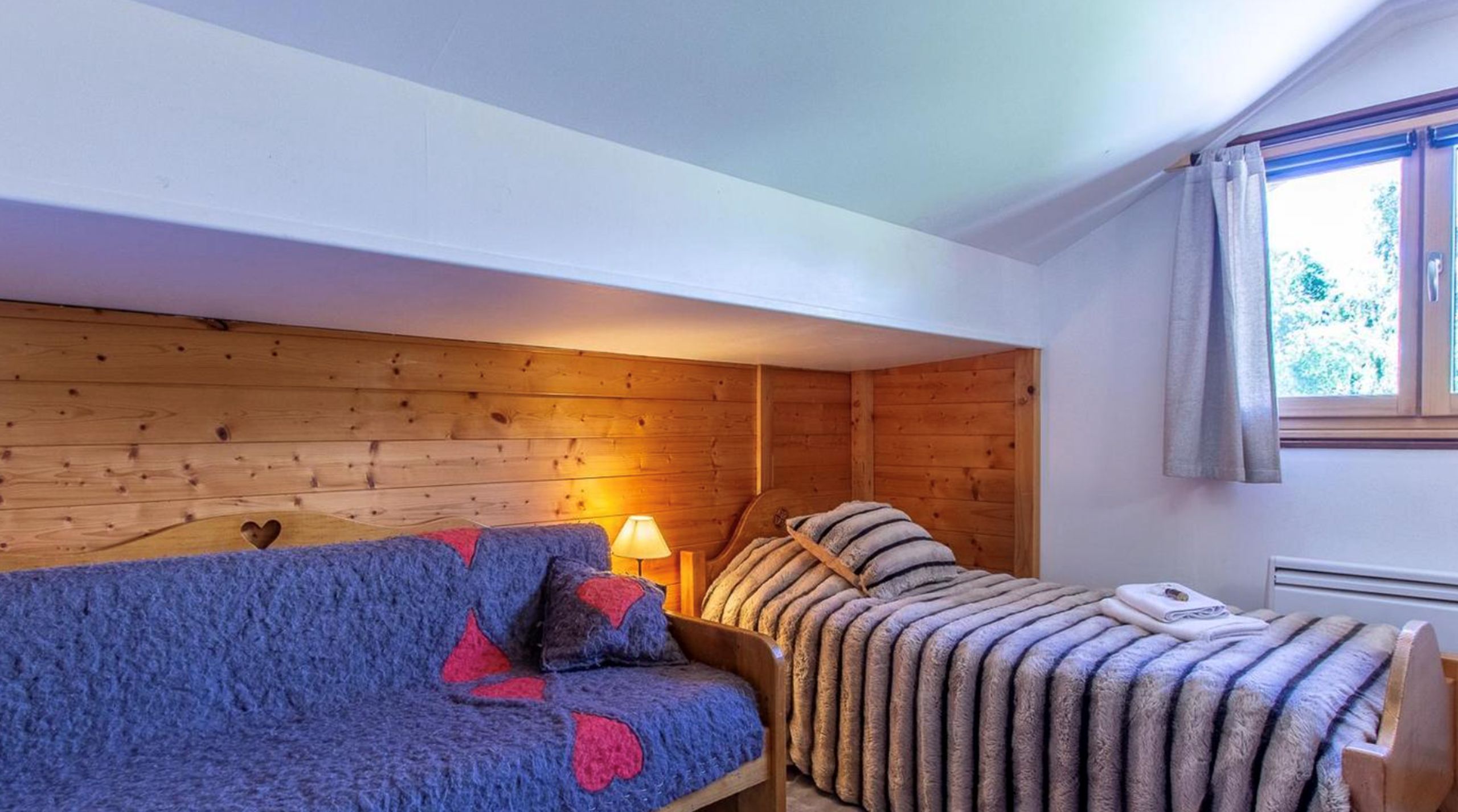 Ein gemütliches Zimmer mit Holzboden, einem blauen Sofa mit roten Akzenten, einem gestreiften Bett und einem Fenster mit Vorhängen.