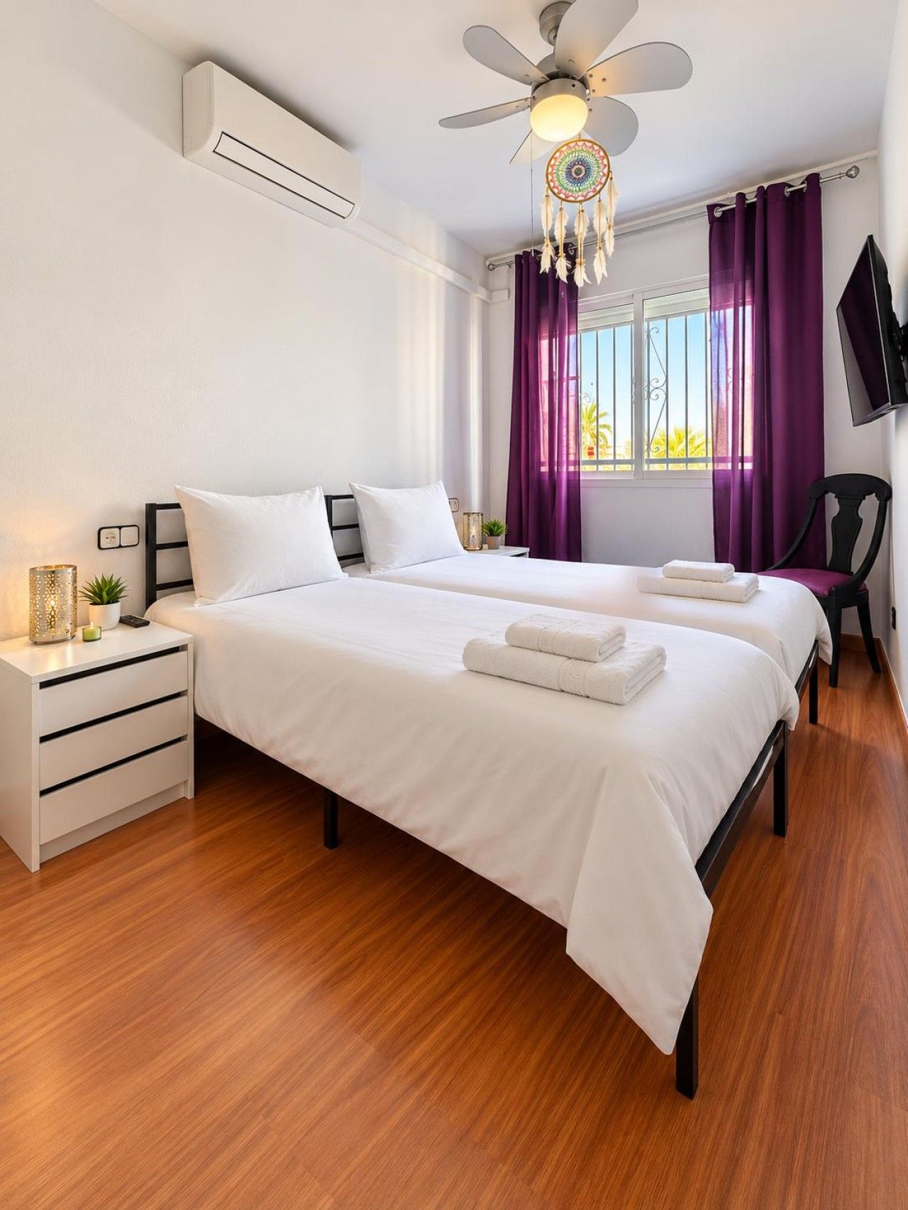 A modern hotel room with two beds, a ceiling fan, a TV, and a window with purple curtains.