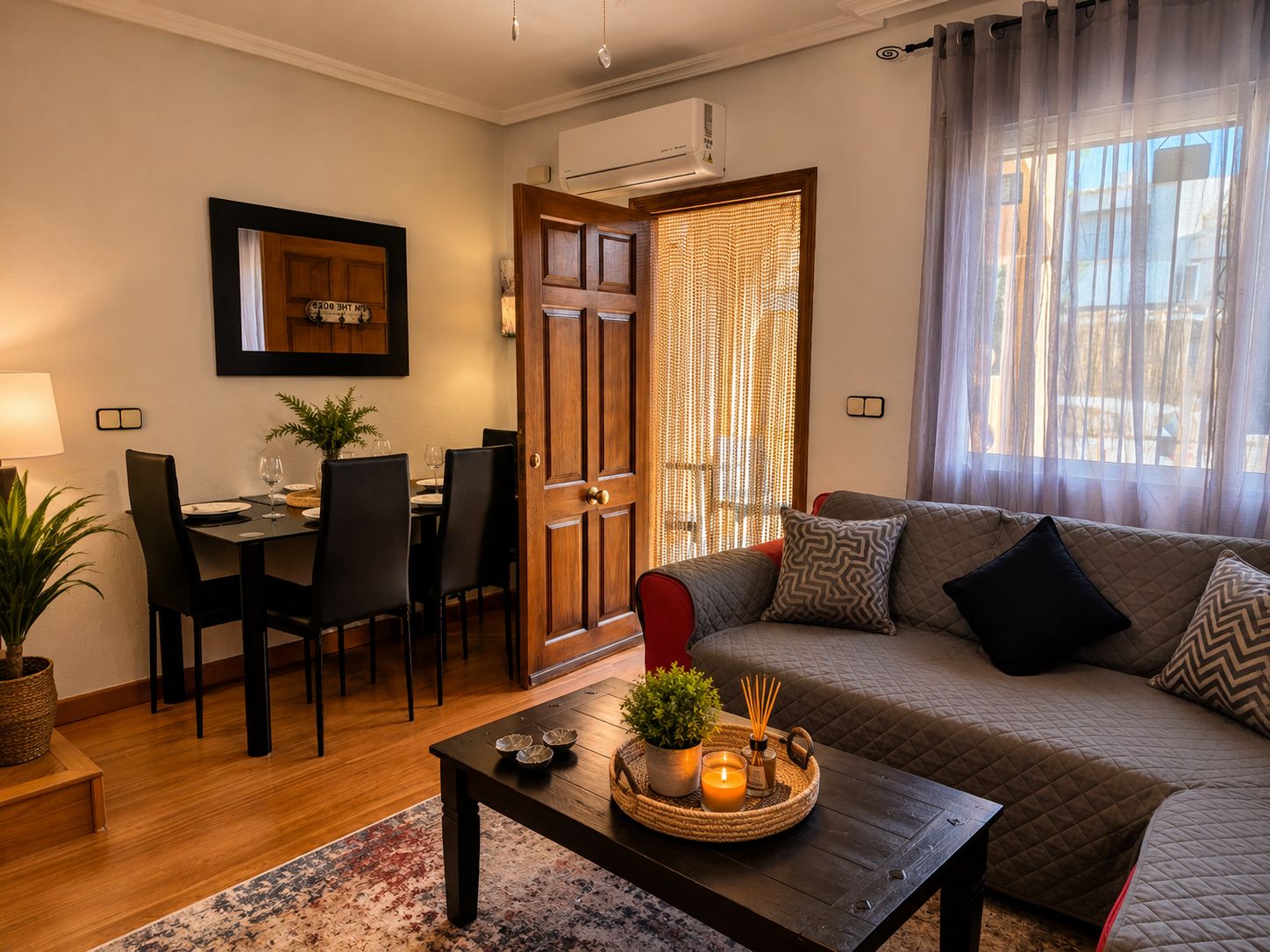A living room with a sofa, a dining table with chairs, a coffee table with decorative items, and a large window with curtains.