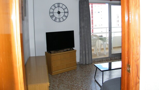 A room with a television on a wooden dresser, a clock on the wall, and a balcony with a table and chairs. - Family apartment, sea view, pets welcome - 6 guests (Gandia)