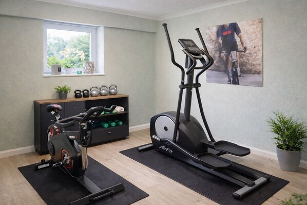 Un gimnasio en casa con una elíptica, una bicicleta estática y una prensa de pesas. Hay una ventana con plantas en el alféizar, un estante con mancuernas y un cartel en la pared.