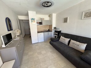 Ein kompakter Wohnbereich mit Küchenzeile, einem schwarzen Sofa mit Kissen, einem Flachbildfernseher und einem kleinen Kühlschrank.