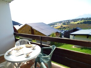 A balcony with a table and chairs, overlooking a rural landscape with houses and mountains. - Charming studio, mountain view - 3 guests (Les Saisies)