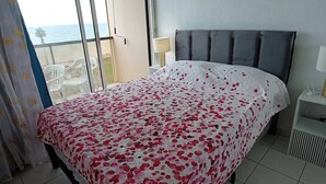 A hotel room with a bed, a nightstand, a lamp, and a view of the sea through a large window. - T2 sea view, parking - 4 guests (Canet-Plage)