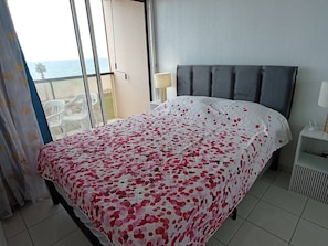 Ein Hotelzimmer mit Bett, Nachttisch, Lampe und Meerblick durch ein großes Fenster.