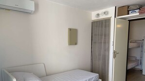 Ein Zimmer mit einem Einzelbett, einem kleinen Nachttisch, einem Spiegel, einem Kleiderschrank und einem Deckenventilator.