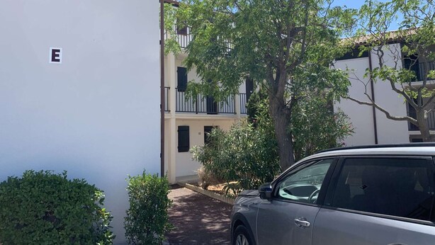 Ein Wohngebäude mit weißer Fassade, ein Baum im Vordergrund und ein geparktes Auto.