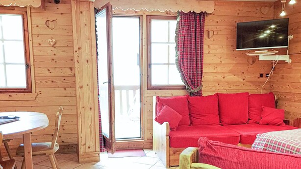Ein Holzblockhaus-Interieur mit einem roten Sofa, einem Holzbett mit herzförmigem Kopfteil, einem an der Wand montierten Fernseher und einem Esstisch mit Stühlen.