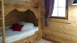 Ein Holzbett mit Etagen, ein rotes Kissen, ein Fenster mit Karomuster-Vorhang und eine Holzbalkendecke.