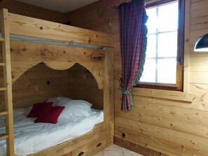 Ein Holzbett mit Etagen, ein rotes Kissen, ein Fenster mit Karomuster-Vorhang und eine Holzbalkendecke.