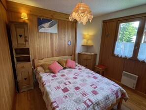 A bedroom with a wooden headboard, a bed with a patterned quilt, a bedside table with a lamp, and a window with curtains. - Cozy apartment, panoramic view - 6 guests (Les Saisies)