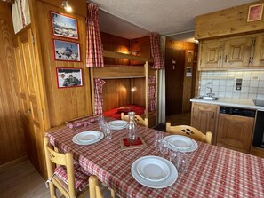 A cozy kitchen with wooden cabinets, a dining table set with plates and glasses, and a bunk bed with plaid curtains. - Cozy apartment, panoramic view - 6 guests (Les Saisies)