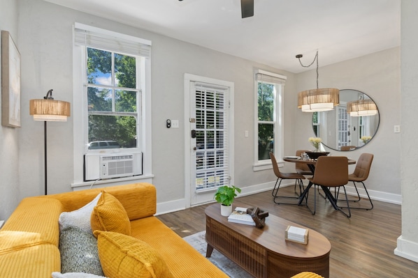 A modern living room with a yellow sofa, a dining area with a table and chairs, and large windows with blinds.