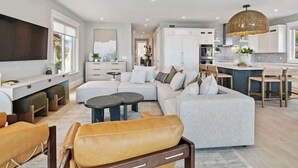 A spacious living room with a large sofa, a coffee table, and a television. There is a kitchen area with white cabinets and a dining space with wooden chairs.