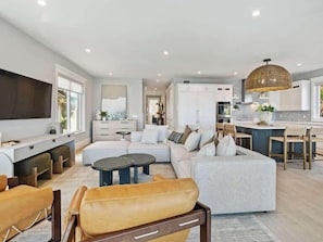 A spacious living room with a large sofa, a coffee table, and a television. There is a kitchen area with white cabinets and a dining space with wooden chairs.