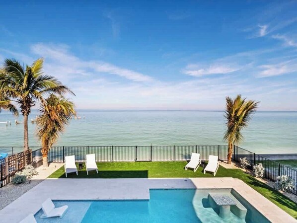 A pool area with a view of the sea, surrounded by palm trees and white lounge chairs.