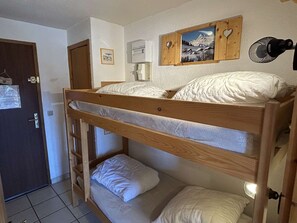Ein Etagenbett mit Holzrahmen, weißer Bettwäsche, einem kleinen Nachttisch, einem an der Wand befestigten Heizgerät und einem Bild von einer Berglandschaft.