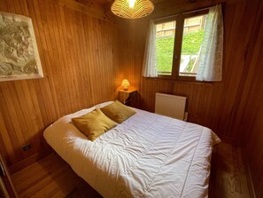 A room with a bed, a nightstand, a lamp, a radiator, and a window with curtains. - Duplex panoramic mountain view - 8 guests (Les Saisies)