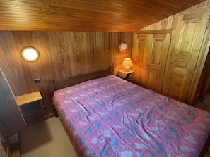 A bedroom with a wooden ceiling, a bed with a patterned bedspread, a nightstand with a lamp, and wooden wardrobe doors.