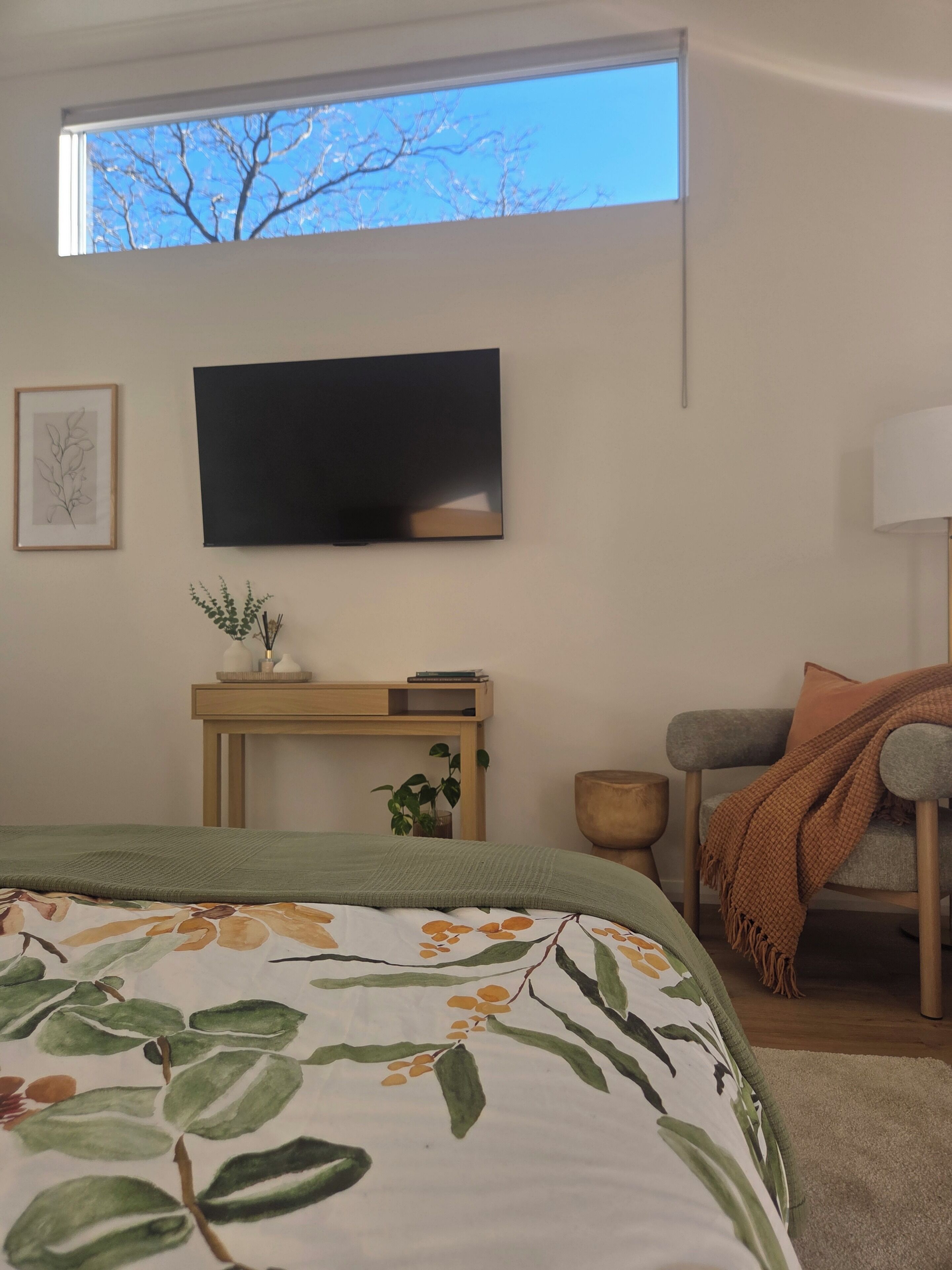 Ein Schlafzimmer mit einem Bett, einem an der Wand montierten Fernseher, einem Nachttisch mit einer Pflanze und Blick auf den Himmel durch ein Fenster.
