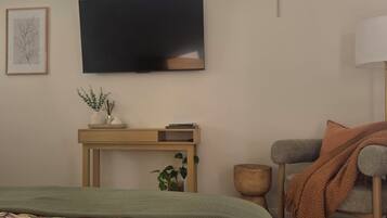 Ein Schlafzimmer mit einem Bett, einem an der Wand montierten Fernseher, einem Nachttisch mit einer Pflanze und Blick auf den Himmel durch ein Fenster.