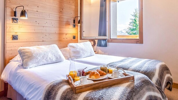 A cozy room with a wooden ceiling, a bed with white bedding, a window with a view of trees, and a tray with breakfast items. - Apartment with panoramic view - 6 guests (Les Saisies)
