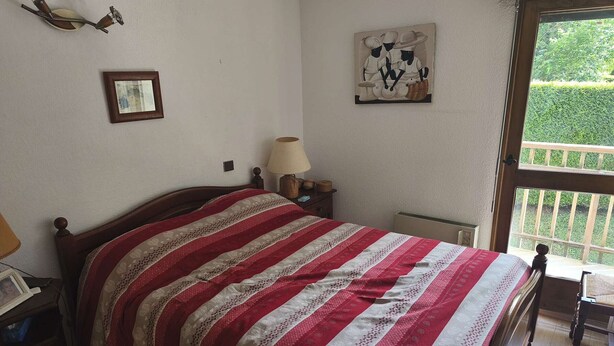 A bedroom with a wooden bed, a bedside table with a lamp, a framed picture on the wall, and a window with a view of greenery. - Apartment with ski view balcony - 4 guests (Arâches-la-Frasse)