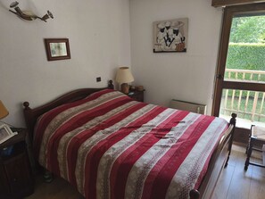 A bedroom with a wooden bed, a bedside table with a lamp, a framed picture on the wall, and a window with a view of greenery. - Apartment with ski view balcony - 4 guests (Arâches-la-Frasse)