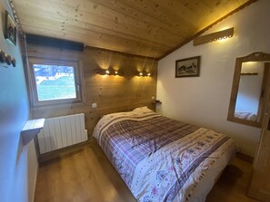 A bedroom with a wooden ceiling, a window, a bed with a patterned bedspread, a bedside table, and a mirror. - Apartment at the foot of the slopes - 8 guests (Valmorel)