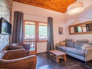 A living room with a wooden ceiling, a television mounted on a stone wall, a sofa, a coffee table, and a view through a glass door. - Duplex with slope view - Valmorel - 6 guests (Valmorel)