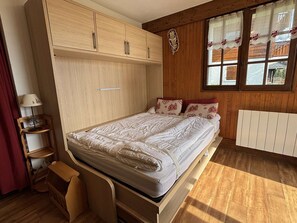A bedroom with a wooden bed, a bedside table, a lamp, and a window with curtains. - Cozy apartment, parking - 4 guests (Les Saisies)