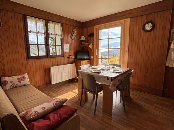 A cozy room with a wooden interior, a dining table set for two, a sofa with patterned cushions, and a view of mountains through the windows. - Cozy apartment, parking - 4 guests (Les Saisies)