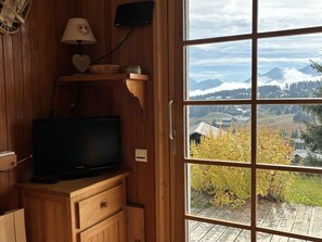 Ein Holzinterieur mit einem Fernseher auf einer Holzkommode, einem Fenster mit Blick auf Berge und Wald, sowie einem an der Wand montierten Lautsprecher.