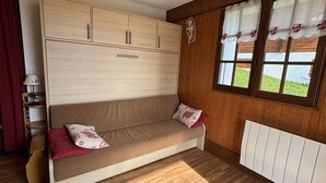 A room with a wooden floor, a beige sofa with red cushions, a wooden wardrobe, a window with curtains, and a radiator. - Cozy apartment, parking - 4 guests (Les Saisies)