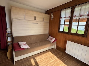 Ein Zimmer mit Holzboden, einem beigen Sofa mit roten Kissen, einem Holzschrank, einem Fenster mit Vorhängen und einem Heizkörper.