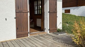 A wooden door with a sliding mechanism, a white exterior wall, and a wooden deck leading to a room with a window and curtains. - Cozy apartment, parking - 4 guests (Les Saisies)