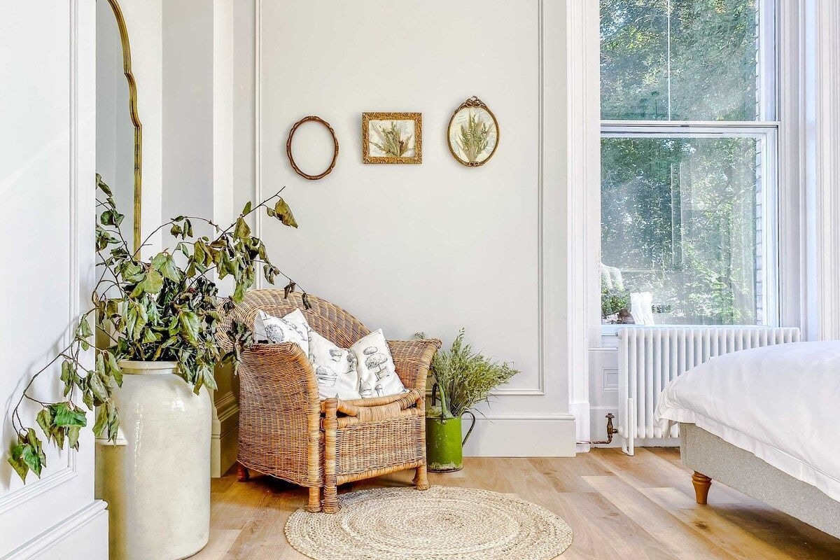 A room with a wicker chair, a large vase with greenery, a round rug, and a bed.