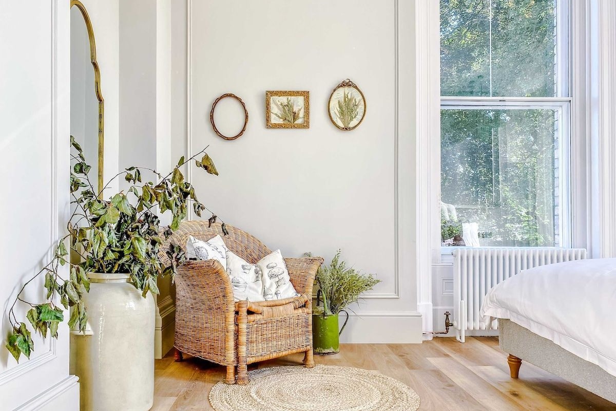 A room with a wicker chair, a large vase with greenery, a round rug, and a bed with white bedding.