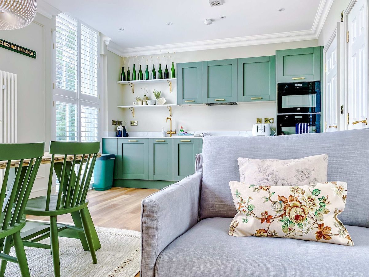 A modern kitchen with teal cabinets, a grey sofa with floral cushions, and a dining area with green chairs.