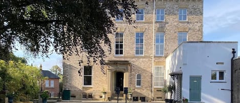 A courtyard with a white table and chairs, surrounded by gravel and grass, in front of a stone building with multiple windows and a green door.