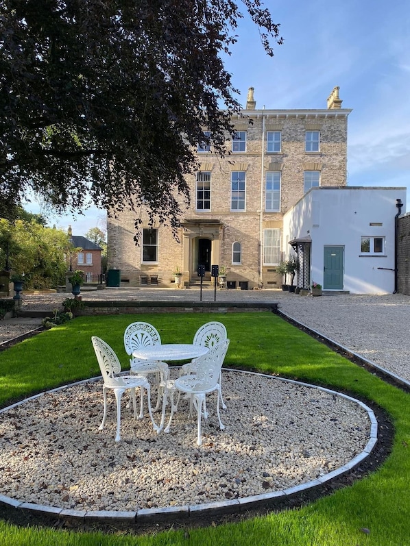 A courtyard with a white table and chairs, surrounded by gravel and grass, in front of a stone building with multiple windows and a green door.